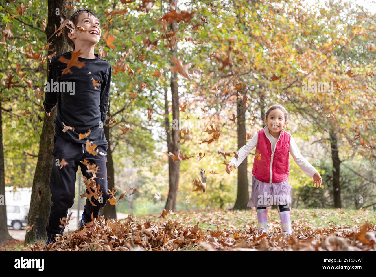 Zwei fröhliche Kinder spielen an einem Herbstnachmittag in einem Park und werfen trockene Blätter in die Luft, umgeben von lebendigen Bäumen Stockfoto