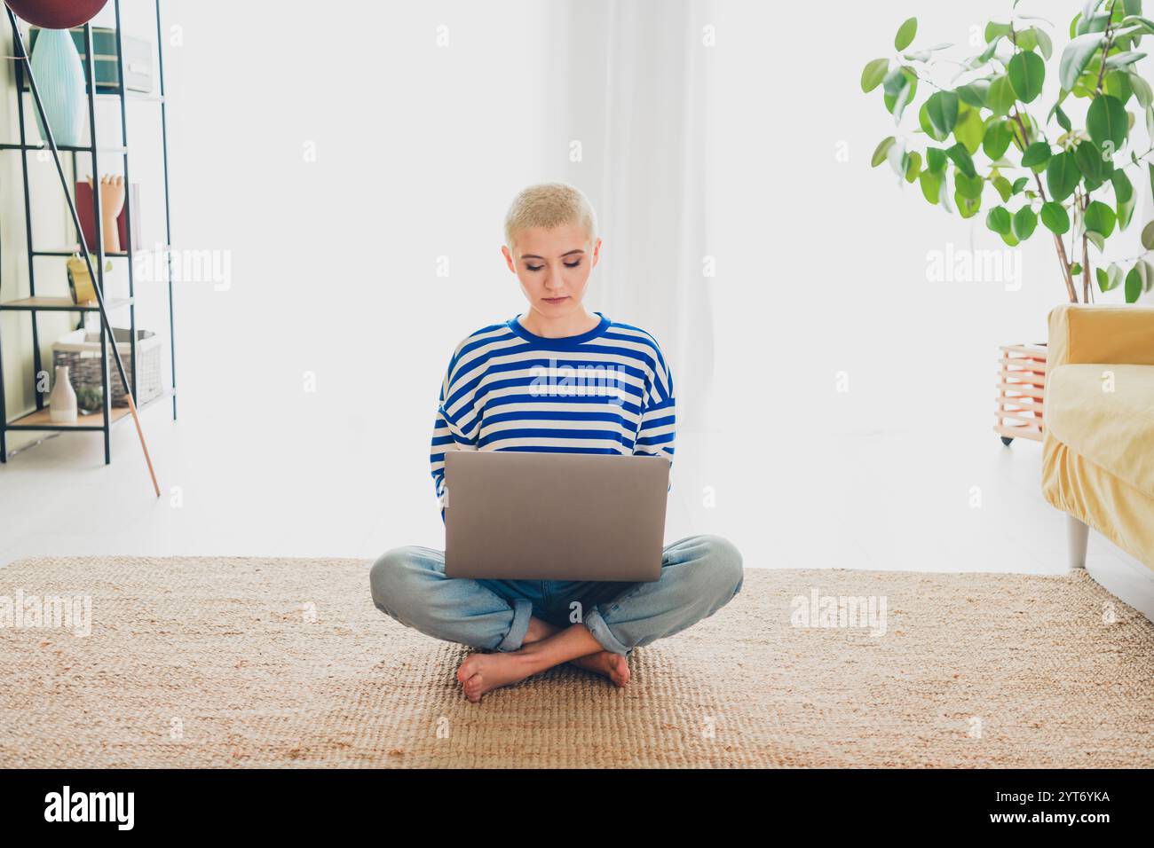 Junge Frau in gestreiftem Pullover, die in entspannter Atmosphäre am Laptop arbeitet und bequem in einem gut beleuchteten Wohnzimmer sitzt Stockfoto