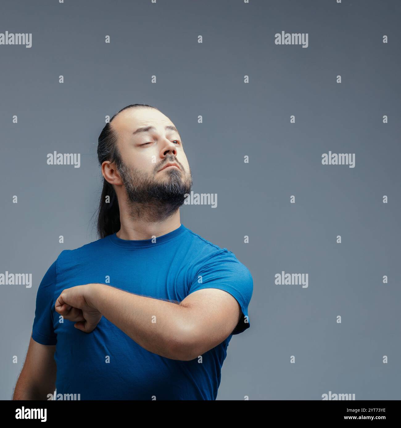 Arroganter junger Mann mit Bart und langen Haaren, ein blaues T-Shirt, schlug sich wie tarzan auf die Brust und strahlt Stolz und Selbstbewusstsein gegen ein Grau aus Stockfoto