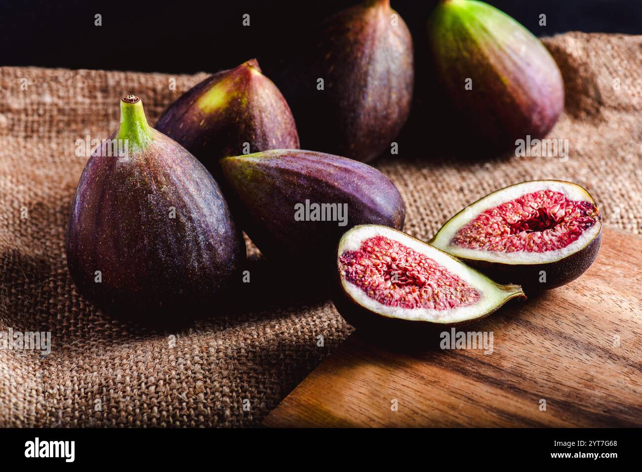 Reife Feigen auf einem rustikalen Sackleinen-Tuch, wobei eine Feige geöffnet wird, um ihr leuchtendes rotes Fleisch und ihre Samen zu enthüllen, die auf einer Holzoberfläche unter weichem Boden liegen Stockfoto
