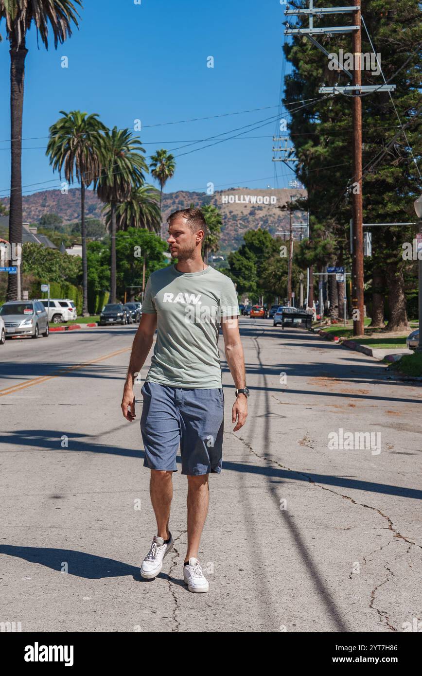 Ein Mann in einem hellgrünen T-Shirt läuft eine palmengesäumte Straße in Los Angeles entlang. Das legendäre Hollywood-Schild ist unter einem klaren blauen Himmel zu sehen, was darauf hindeutet Stockfoto