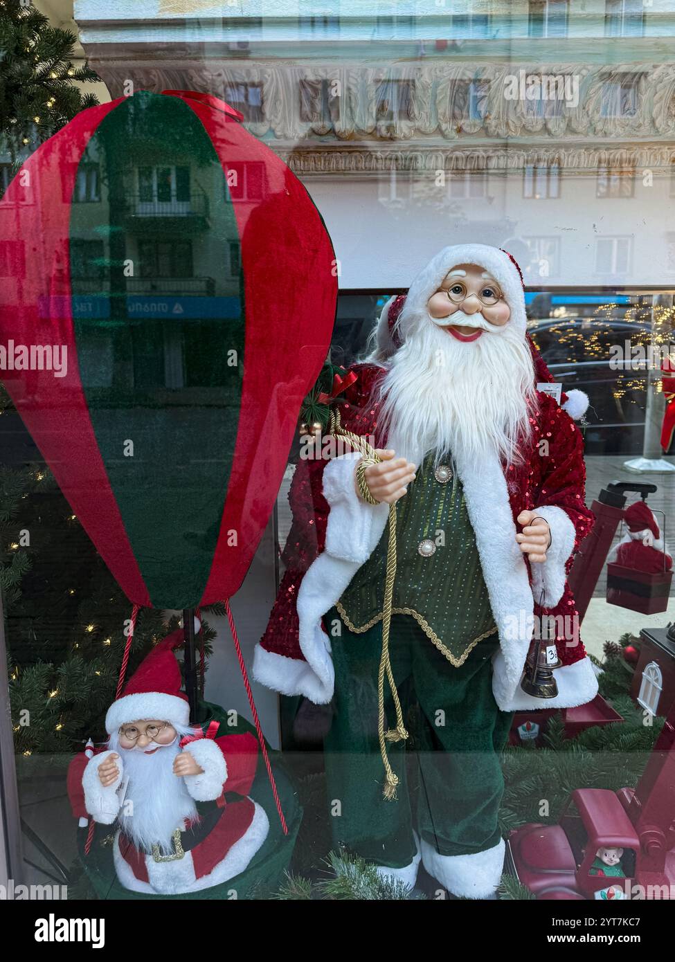 Dieses Foto am Tag zeigt ein festliches Fenster mit zwei Weihnachtsmannsfiguren in klassischer rot-grüner Kleidung. Ein Weihnachtsmann steht aufrecht Stockfoto