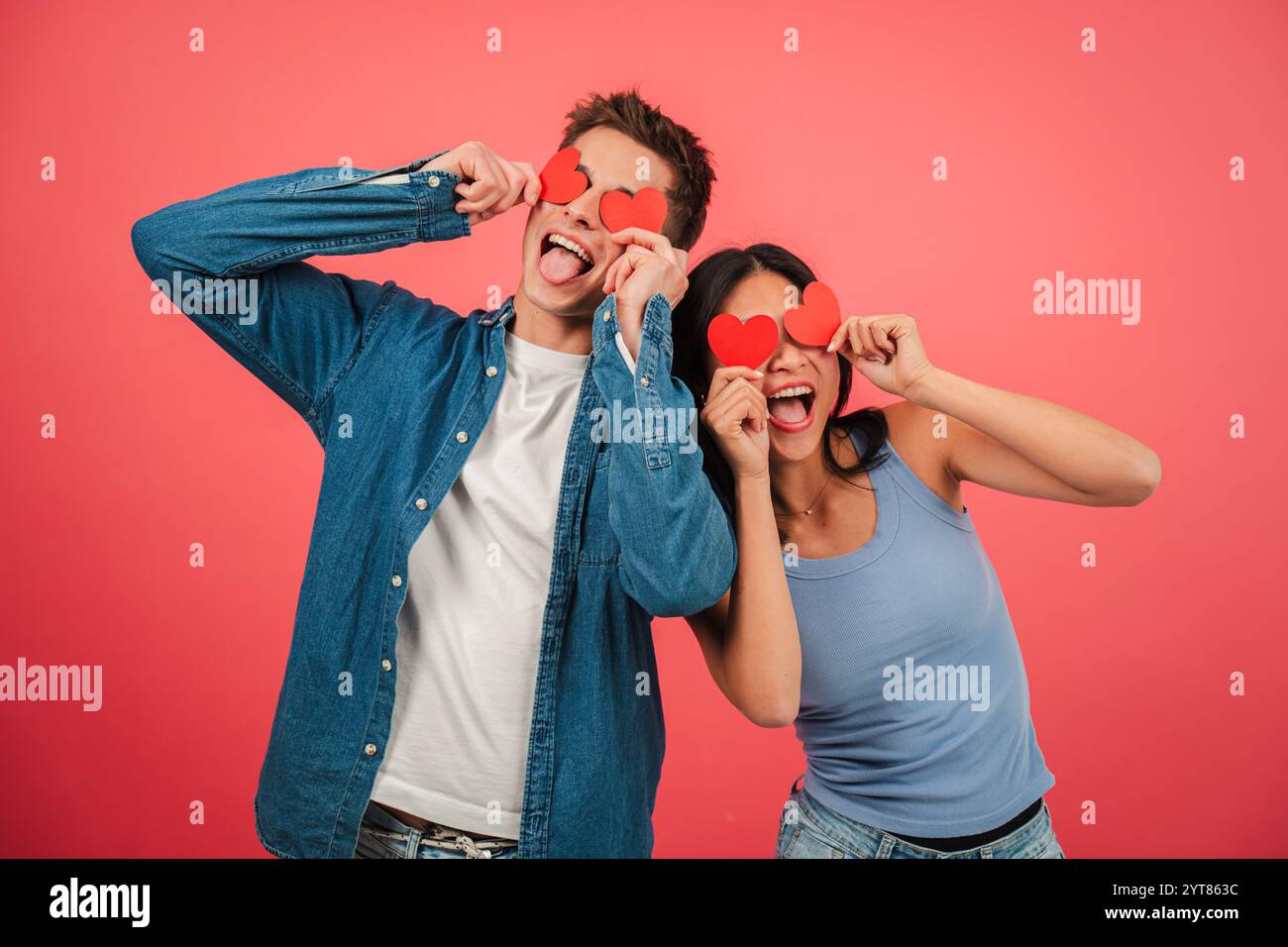 Verspieltes junges Paar mit roten Herzformen und lustigen Gesichtern vor leuchtendem rotem Hintergrund, das Liebe und Glück mit fröhlichen Ausdrücken in einem lustigen und kreativen Valentinstag feiert. Hochwertige Fotos Stockfoto