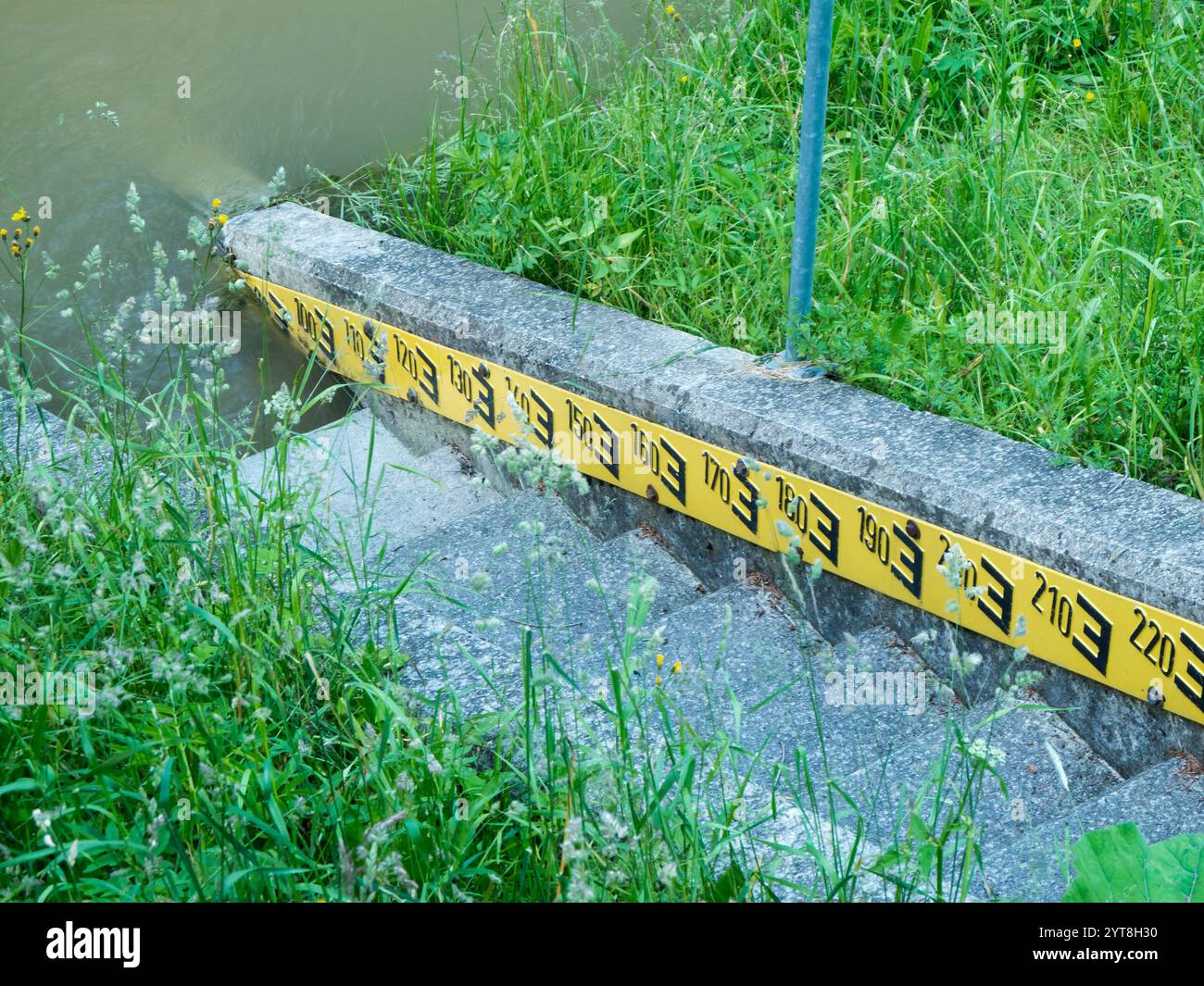 Wasserstandsanzeige mit Steintreppen und schwarzer Schrift auf gelbem Hintergrund auf einem grasgesäumten Damm. Stockfoto