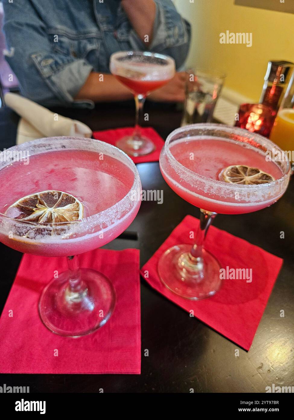 Zwei elegante Gläser rosafarbener Cocktails, garniert mit getrockneten Zitrusscheiben auf knalligen rosa Servietten, mit einer Person im Hintergrund. Stockfoto