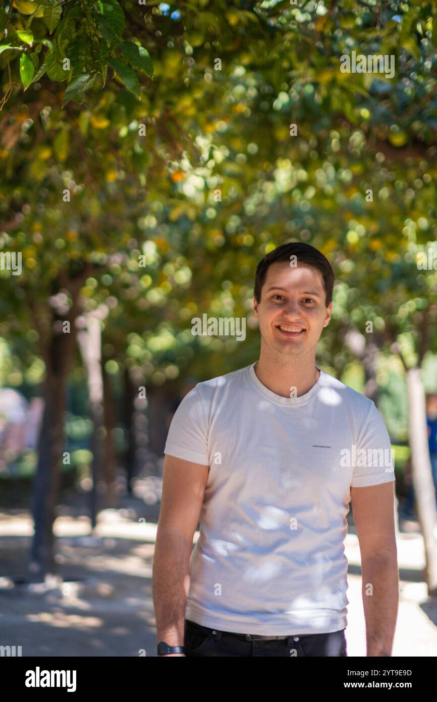 Junger Mann in legeren Sommerbekleidung, der auf einem von Bäumen gesäumten Pfad im mediterranen Garten Malagas spaziert, wobei das Sonnenlicht natürliche Muster erzeugt. Stockfoto