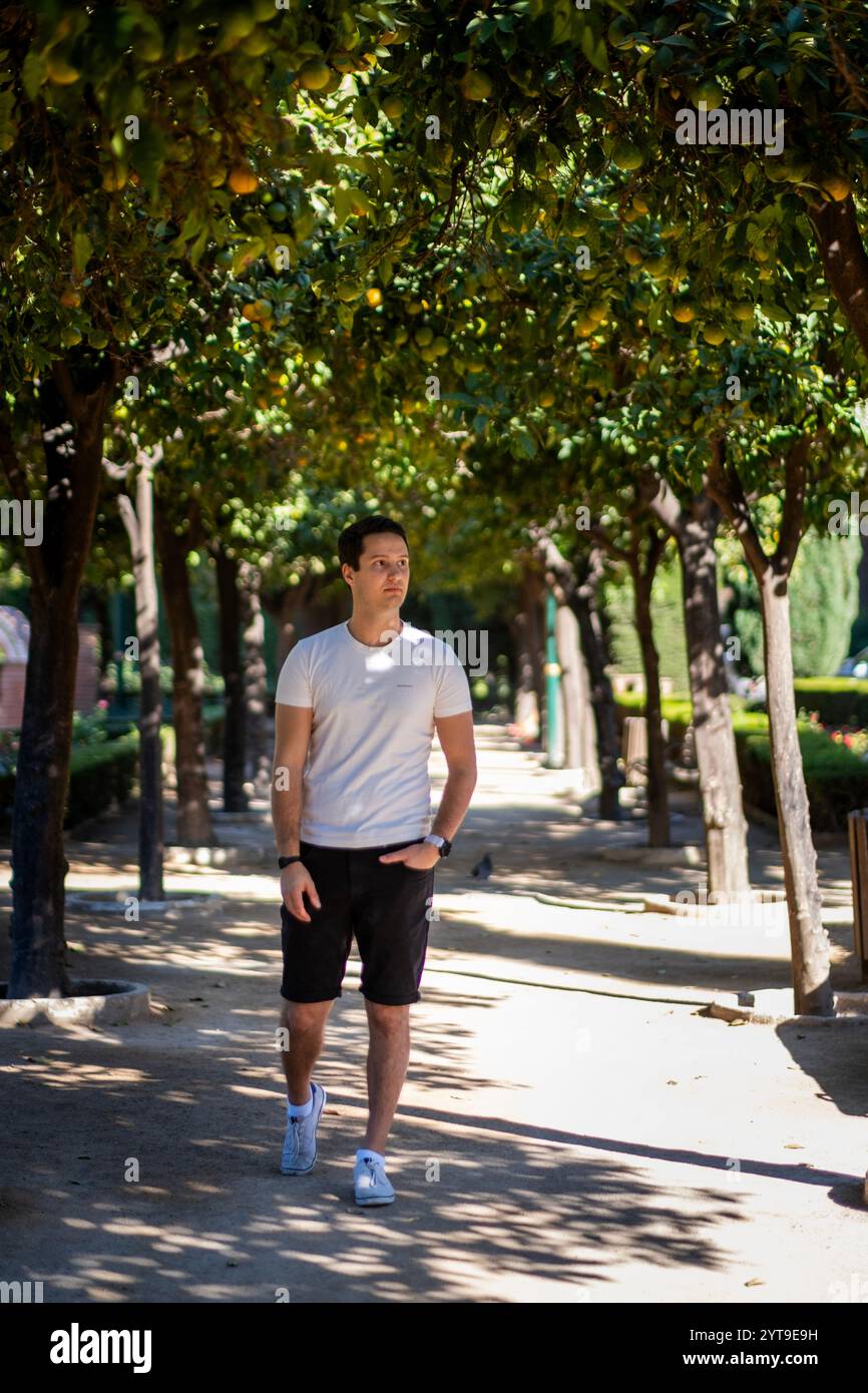 Junger Mann in legeren Sommerbekleidung, der auf einem von Bäumen gesäumten Pfad im mediterranen Garten Malagas spaziert, wobei das Sonnenlicht natürliche Muster erzeugt. Stockfoto