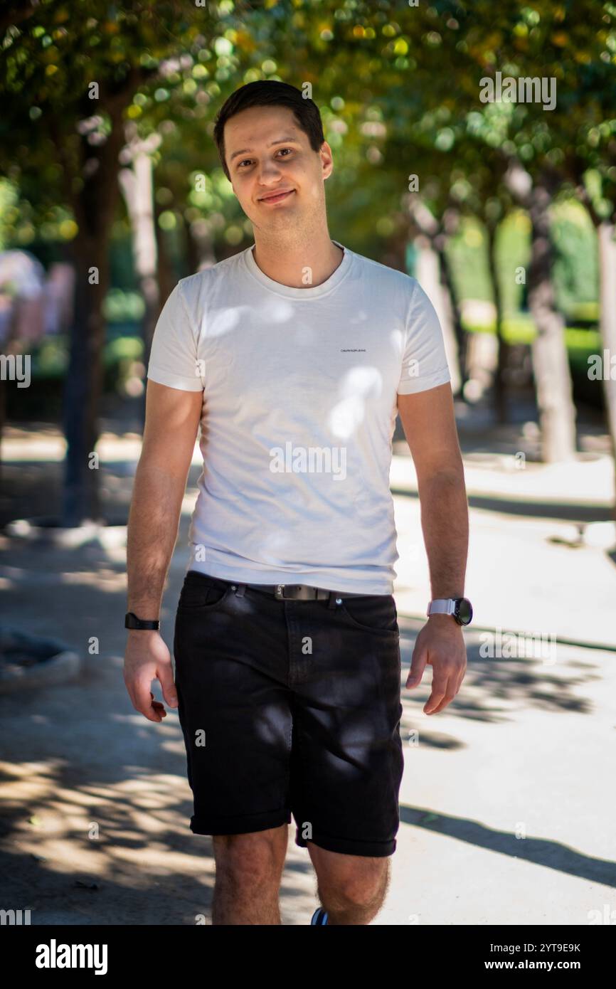 Junger Mann in legeren Sommerbekleidung, der auf einem von Bäumen gesäumten Pfad im mediterranen Garten Malagas spaziert, wobei das Sonnenlicht natürliche Muster erzeugt. Stockfoto