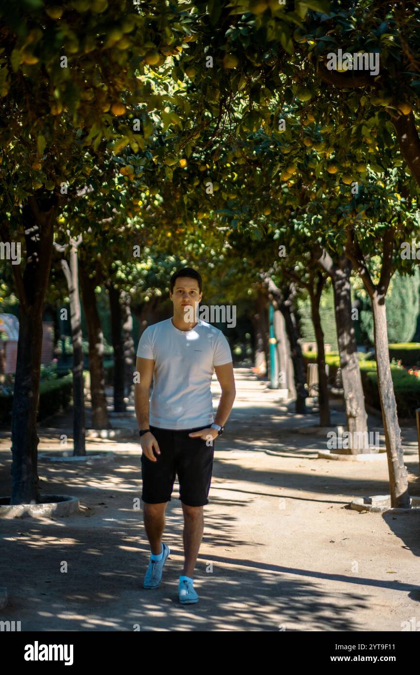 Junger Mann in legeren Sommerbekleidung, der auf einem von Bäumen gesäumten Pfad im mediterranen Garten Malagas spaziert, wobei das Sonnenlicht natürliche Muster erzeugt. Stockfoto