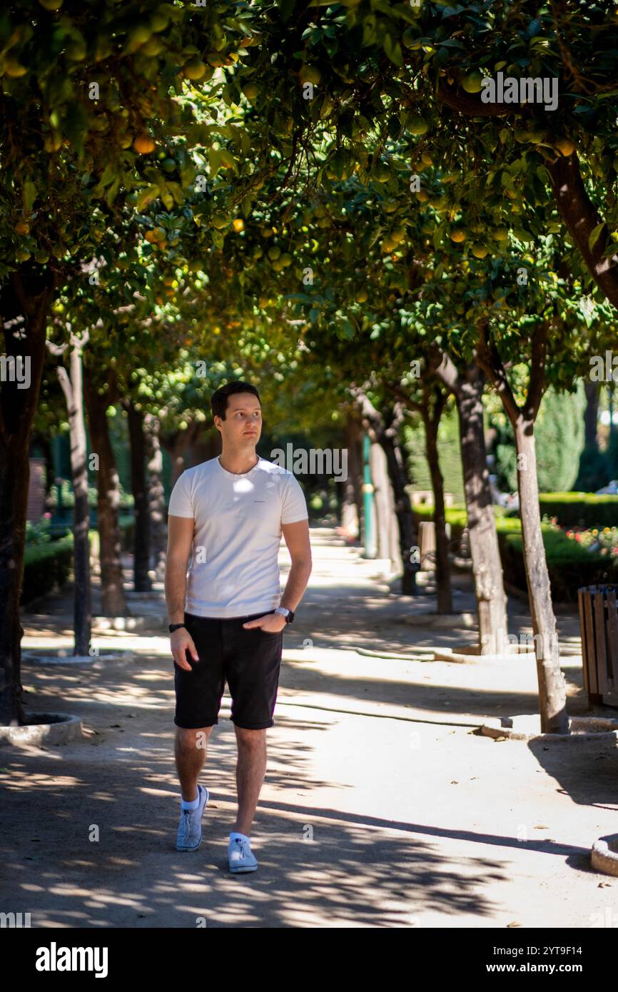 Junger Mann in legeren Sommerbekleidung, der auf einem von Bäumen gesäumten Pfad im mediterranen Garten Malagas spaziert, wobei das Sonnenlicht natürliche Muster erzeugt. Stockfoto