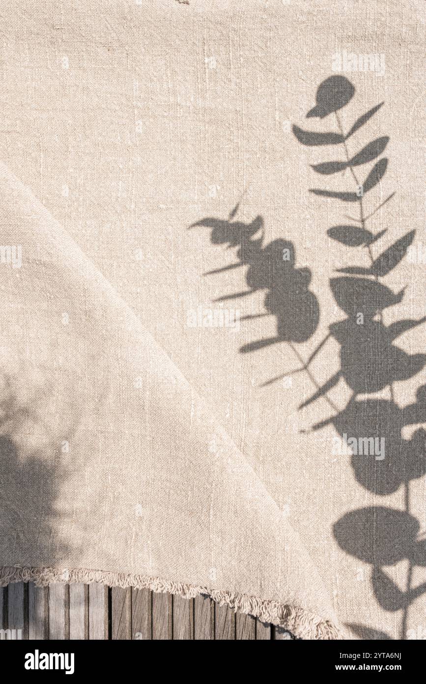 Leinentischdecke im Sonnenlicht mit Blattschatten. Beigefarbene Leinenstoff auf rustikalem Holztisch. Vertikaler Hintergrund für Hochzeits- und Geburtstagsfeiern im Boho-Stil mit Platz für Text. Stockfoto