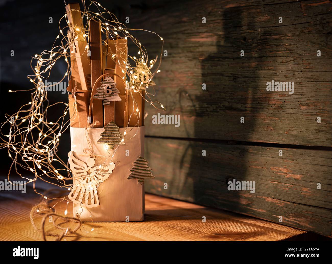 Handgefertigter Adventskalender aus Karton. Kleine Kartonschachteln in einer Papiertüte, verziert mit einer Lichterkette auf rustikalem Holzhintergrund. Umweltfreundliche weihnachtsverpackung. Hintergrund für eine weihnachtsgrußkarte. Stockfoto
