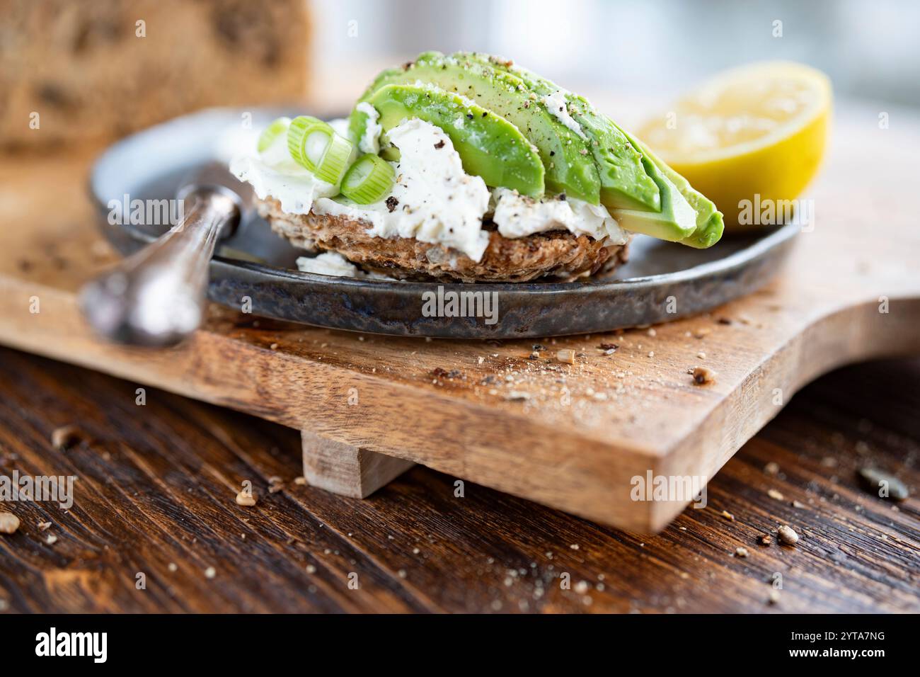 Frisches Mehrkornbrot mit Frischkäse und Avocado auf schwarzer Keramikplatte. Nahaufnahme vor einem hellen Fenster. Kurze Schärfentiefe. Stockfoto