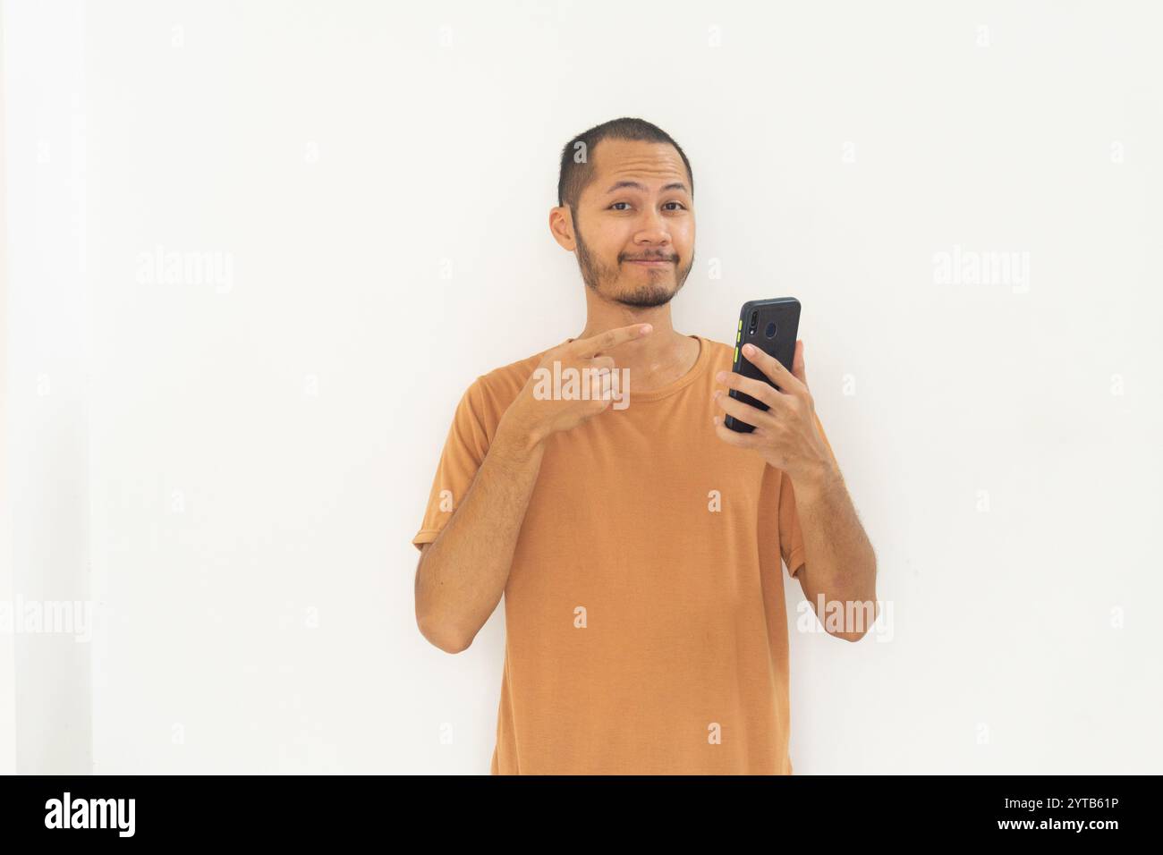 Junge Männer, die ein oranges Hemd tragen, zeigen auf ein lächelndes Handy auf isoliertem weißem Hintergrund Stockfoto
