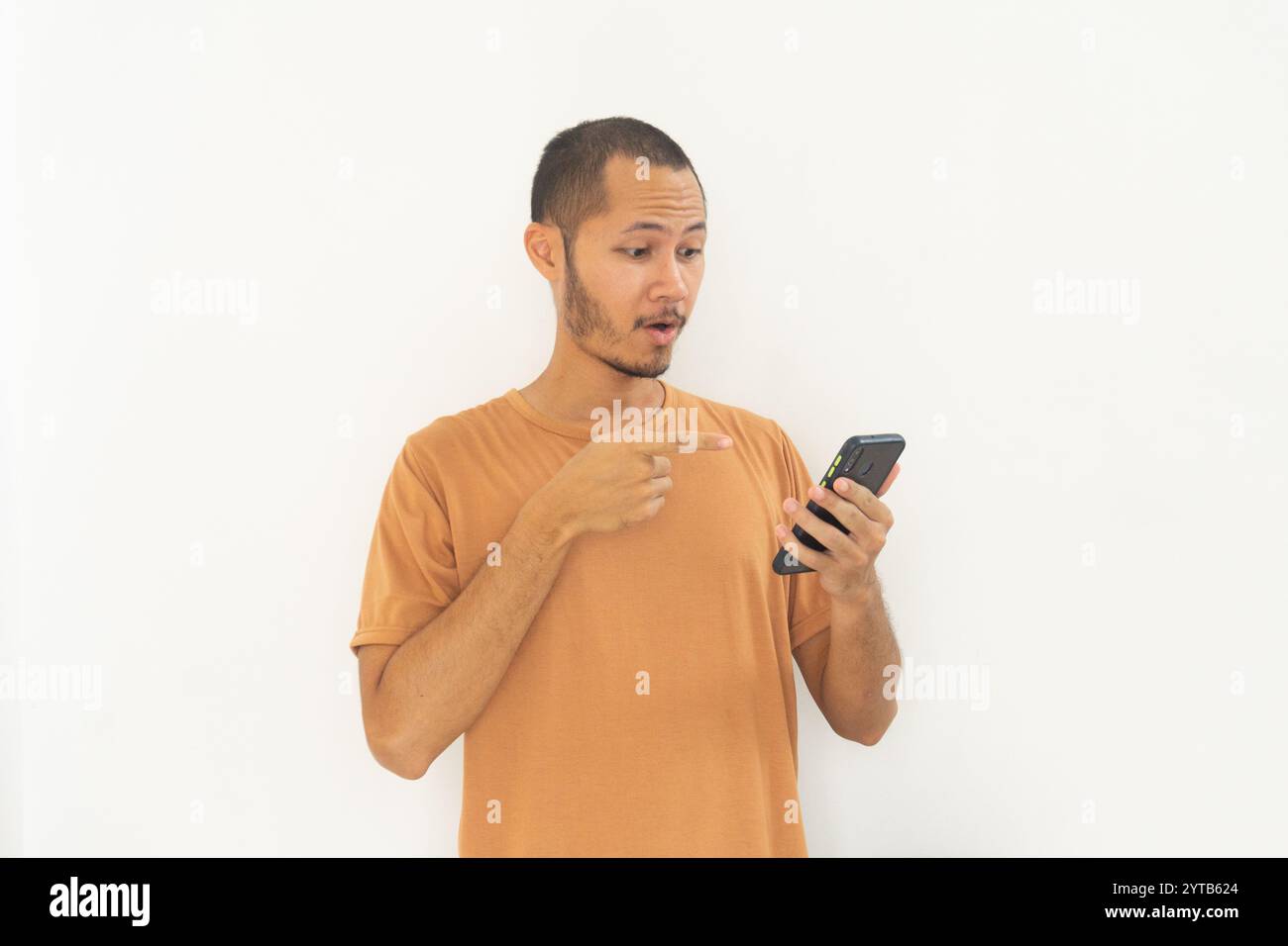 Junge Männer, die ein oranges Hemd tragen, zeigen auf ein lächelndes Handy auf isoliertem weißem Hintergrund Stockfoto