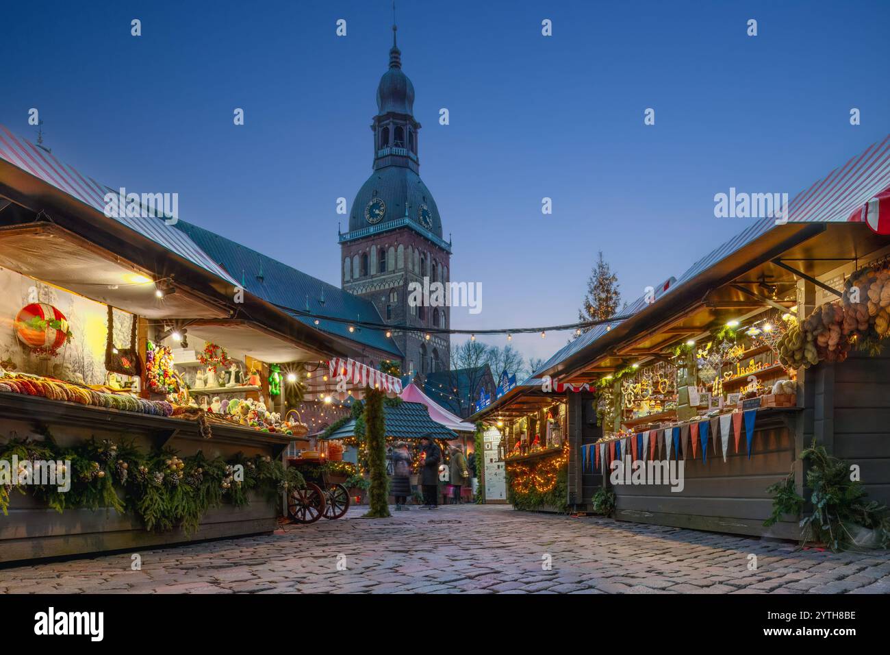 Blick auf einen Weihnachtsmarkt, gesäumt von dekorierten Verkaufsständen und funkelnden Lichtern. Der Dom-Turm und die Bewegung verwischten unbekannte Menschen im Stockfoto