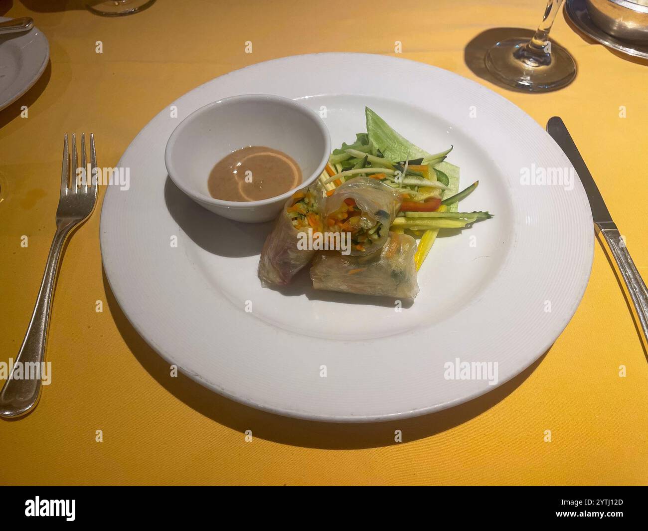 Ein weißer Teller einer Sommerrolle; knuspriges Gemüse in Reispapier mit cremiger Erdnussdippsoße. Das Geschirr wird von Besteck umgeben, zusammen mit einem Glas Stockfoto