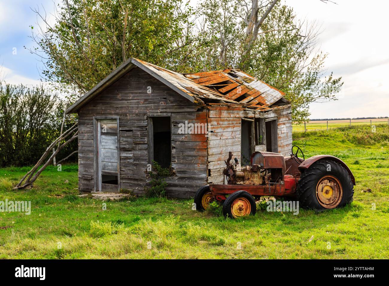 Ein alter roter Traktor steht vor einem baufälligen Haus. Das Haus ist in einer ländlichen Umgebung mit vielen Bäumen im Hintergrund. Das Konzept des Nostals Stockfoto