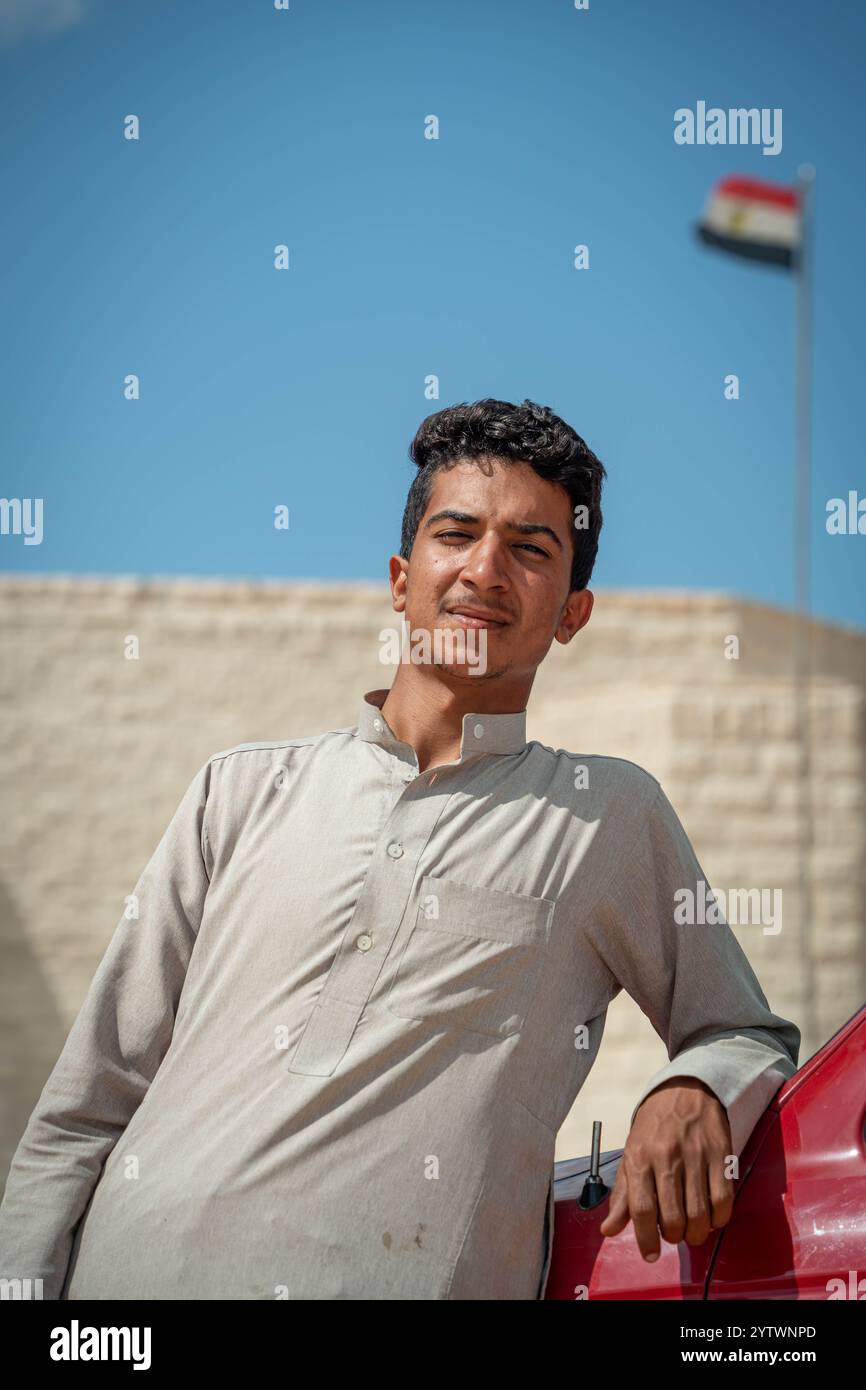 Porträt eines ägyptischen Taxifahrers, der neben seinem Fahrzeug steht, mit der ägyptischen Flagge im Hintergrund auf dem deutschen Kriegsfriedhof El Alamein. Stockfoto