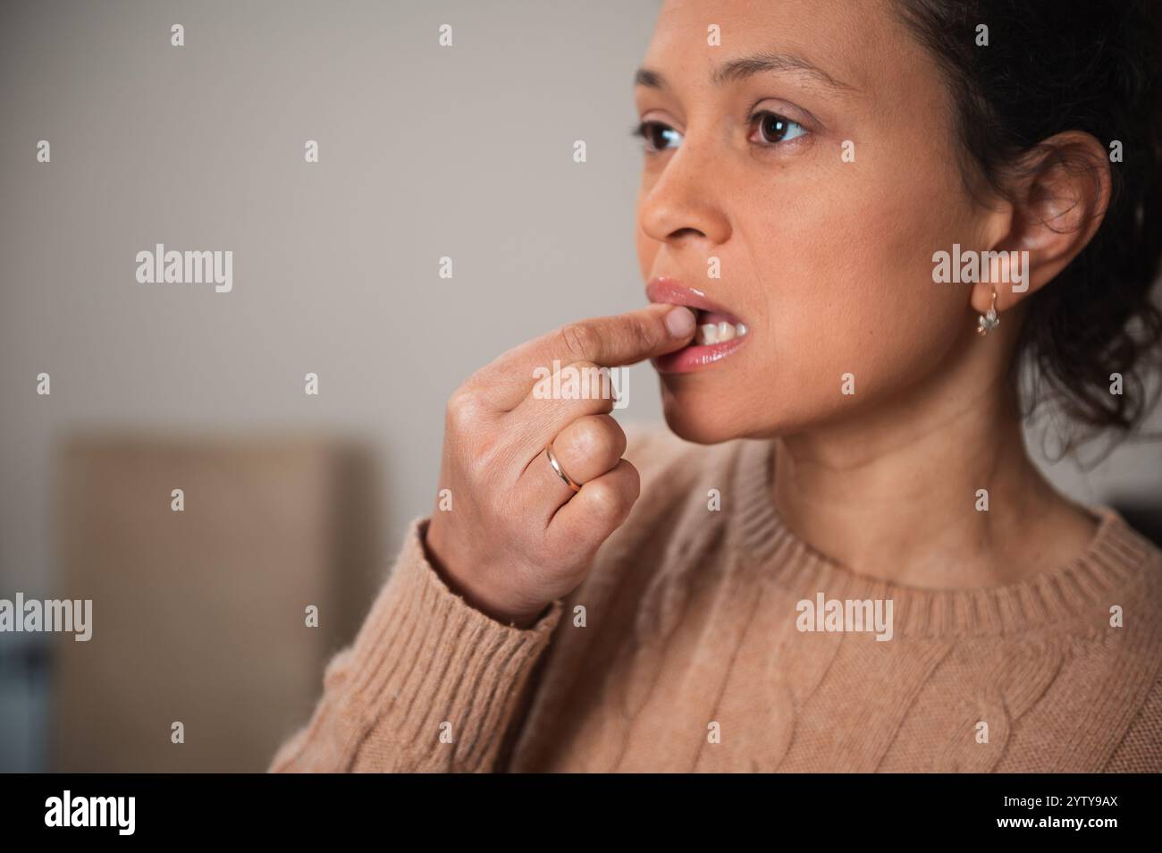 Eine Nahaufnahme einer Frau, die ihre Lippen mit dem Finger untersucht. Sie wirkt konzentriert und nachdenklich, trägt einen beigefarbenen Pullover und Ohrringe, was einen andeutet Stockfoto