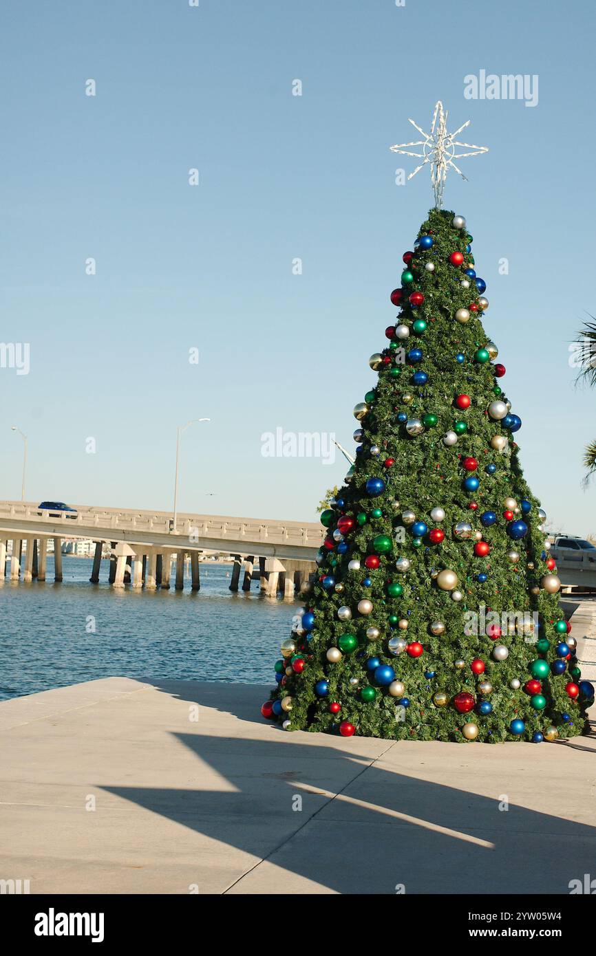 Vertikaler Blick auf einen rot, weiß und grün dekorierten Weihnachtsbaum mit Kugeln. Neben Boca Ciega Bay hinten. In St Pete Beach, FL. Blauer Himmel und ruhiges Wasser. Stockfoto