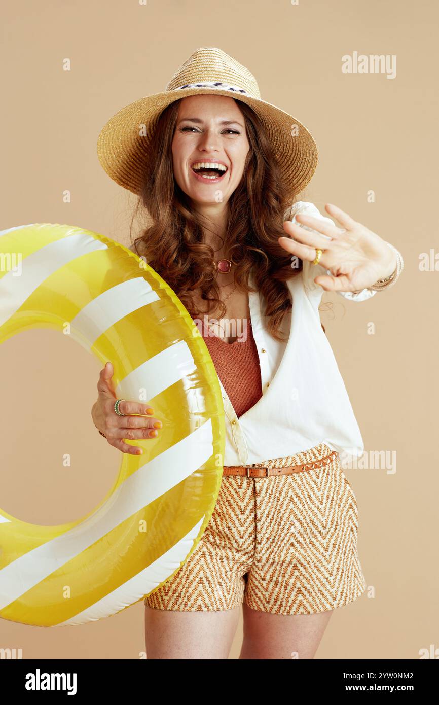 Strandurlaub. Lächelnde moderne 40-jährige Frau in weißer Bluse und Shorts vor beigefarbenem Hintergrund mit aufblasbarem Kreis und Sommerhut. Stockfoto Strandurlaub. Lächelnde moderne 40-jährige Frau in weißer Bluse und Shorts vor beigefarbenem Hintergrund mit aufblasbarem Kreis und Sommerhut. Stockfoto