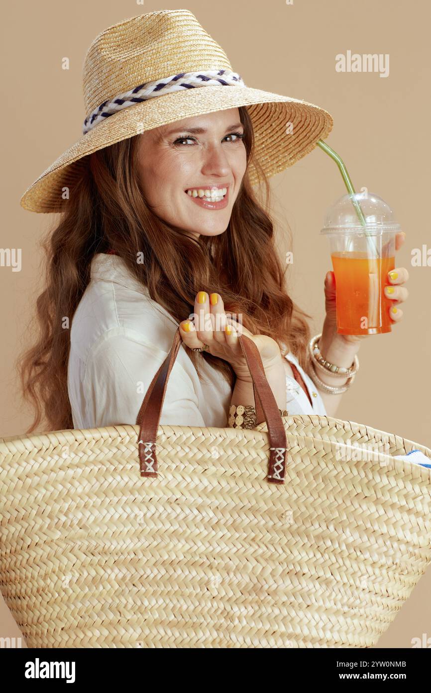 Strandurlaub. Glückliche trendige 40 Jahre alte Hausfrau in weißer Bluse und Shorts isoliert auf Beige mit Strohbeutel, Karotten-Smoothie und Sommerhut. Stockfoto Strandurlaub. Glückliche trendige 40 Jahre alte Hausfrau in weißer Bluse und Shorts isoliert auf Beige mit Strohbeutel, Karotten-Smoothie und Sommerhut. Stockfoto