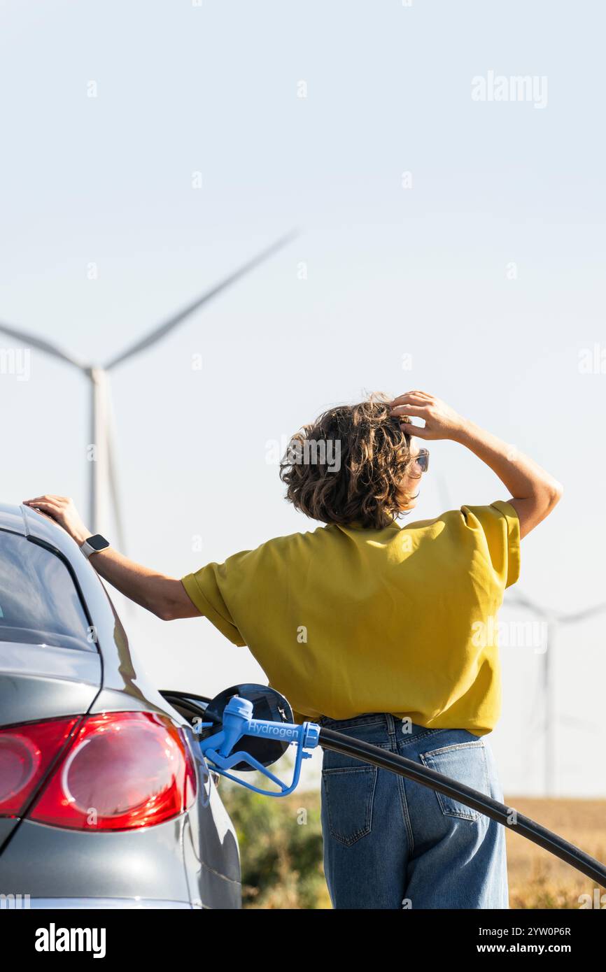 Eine Frau mit einem Wasserstoff-Auto. Betanken des Autos mit Wasserstoffkraftstoff. Windräder im Hintergrund. Stockfoto