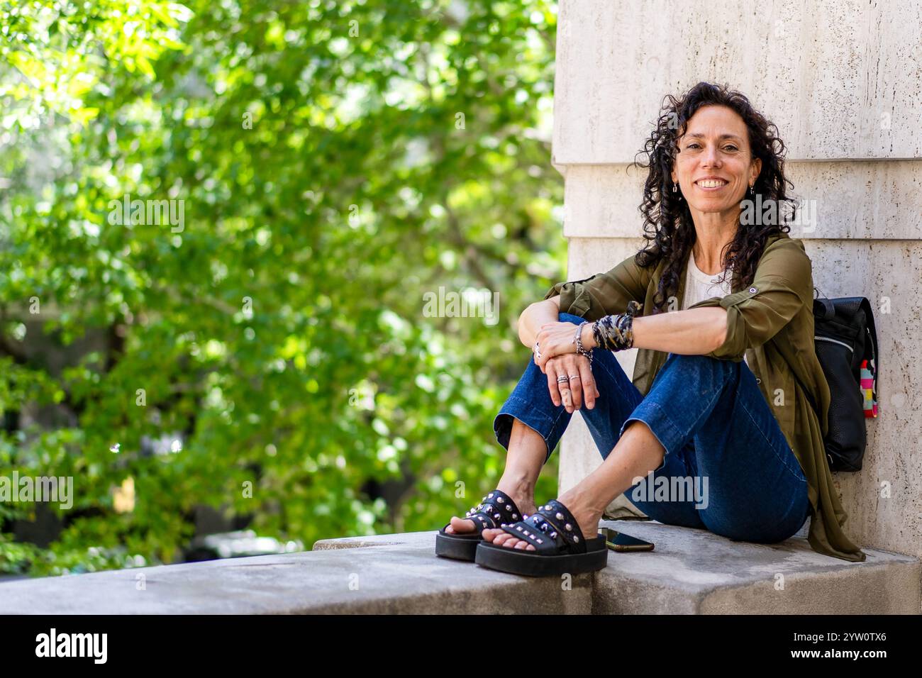 Entspannte Frau im mittleren Erwachsenenalter, die auf einem Felsvorsprung sitzt und die Natur genießt, während sie in die Kamera blickt Stockfoto