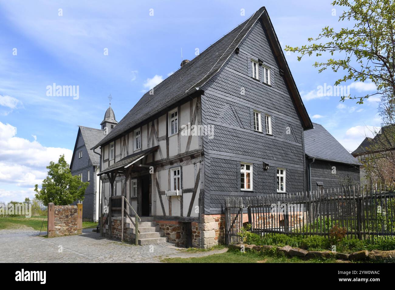 Ein historisches Fachwerkhaus mit Schieferverkleidung, Holzbalken und einem geneigten Dach, umgeben von einem Kopfsteinpflasterweg, Zäunen und üppigem Grün Stockfoto