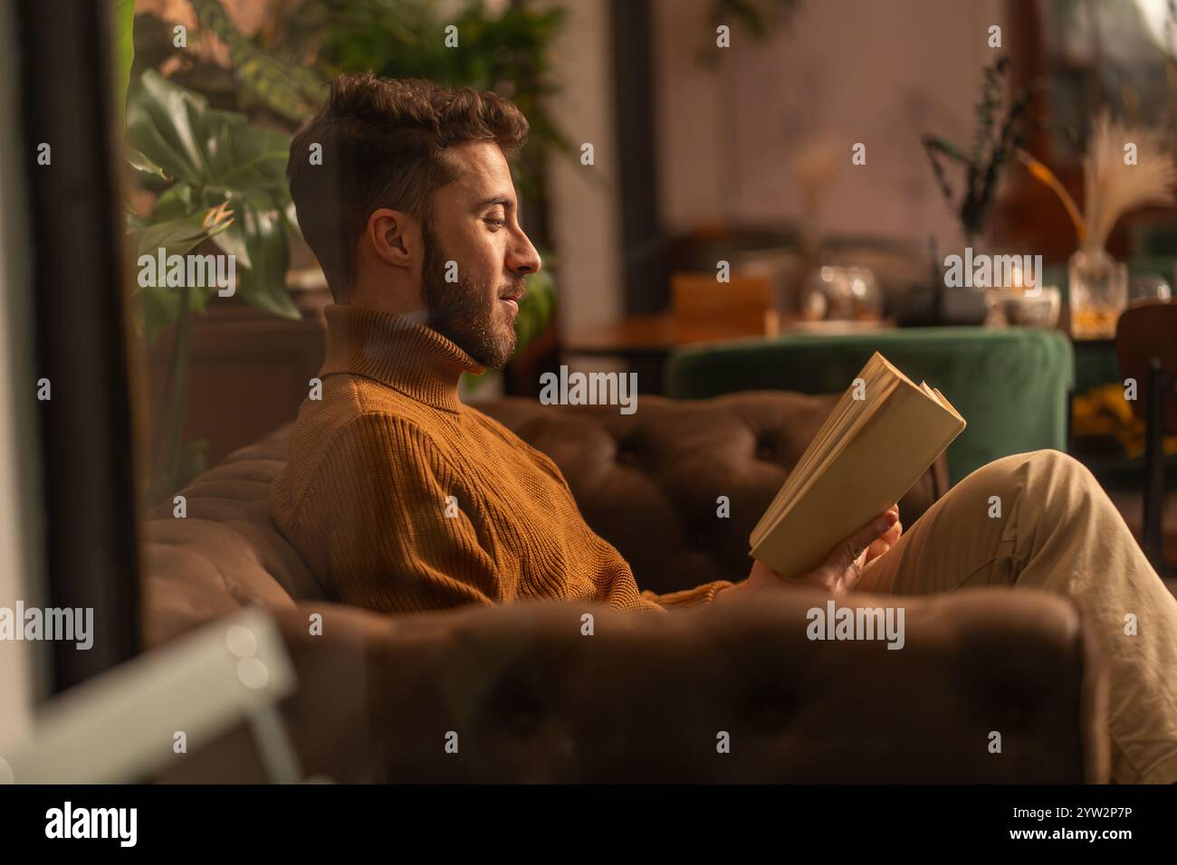 Junger Mann im Rollkragenpullover, der Buch im gemütlichen Café liest, Seitenprofil durch Fenster Stockfoto