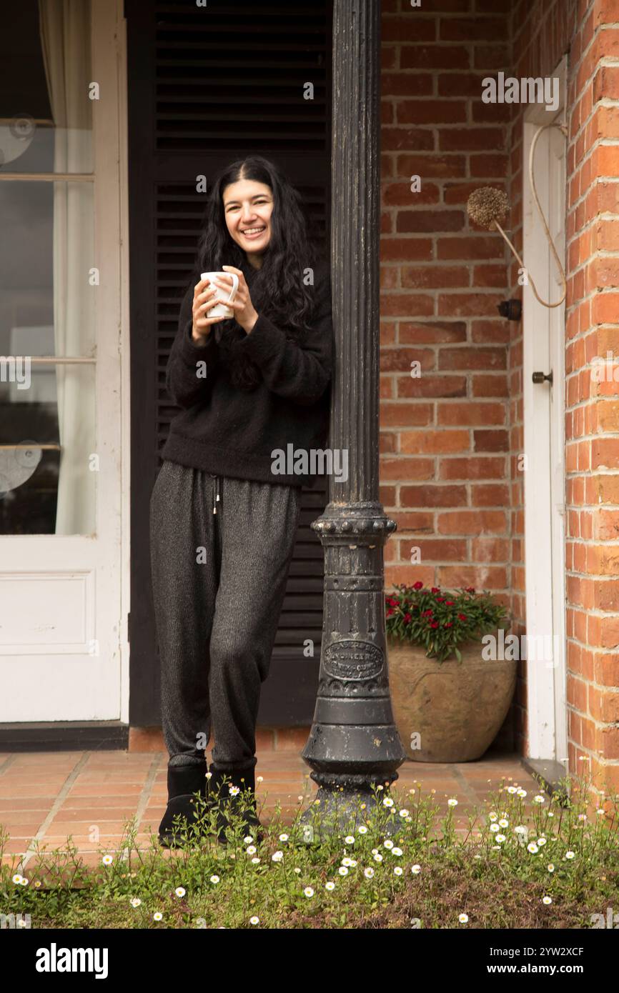Frau, die mit einer Kaffeetasse an einer Tür steht, lächelt und einen gemütlichen Moment im Freien genießt, Auckland, Neuseeland Stockfoto