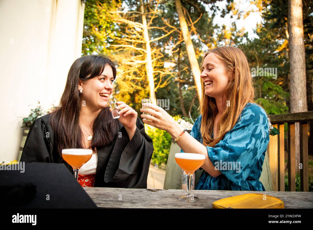 Zwei Frauen, die lachen und mit Gläsern klirren, feiern den Abschlussfeiertag an einem Tisch im Freien, umgeben von Grün, Bournemouth, Dorset UK Stockfoto