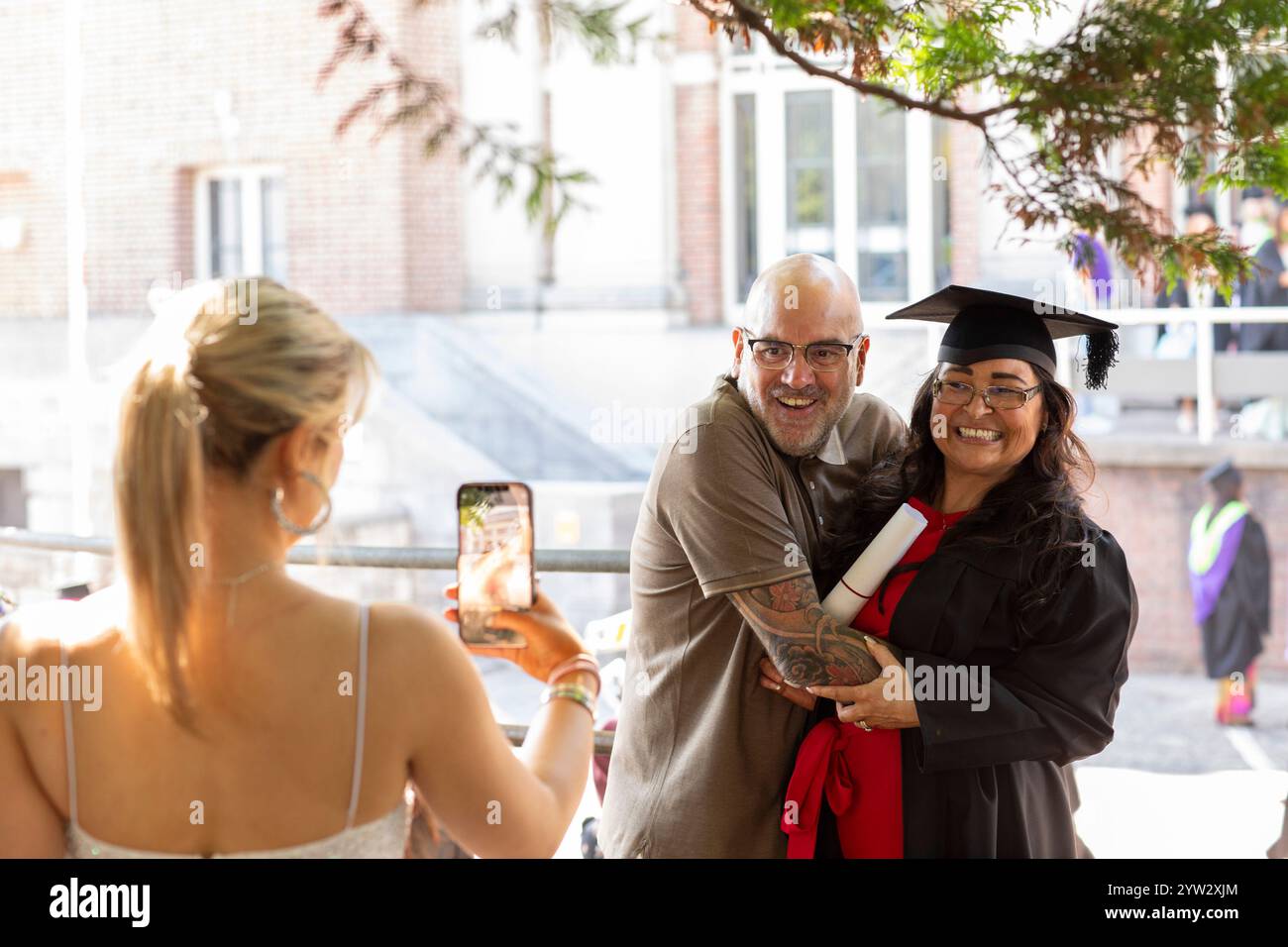 Lächelnde Frau in Abschlussmütze und Kleid posiert für ein Foto mit einem Mann, während eine andere Person ihr Foto mit einem Smartphone macht, Bournemouth, Dorset UK Stockfoto