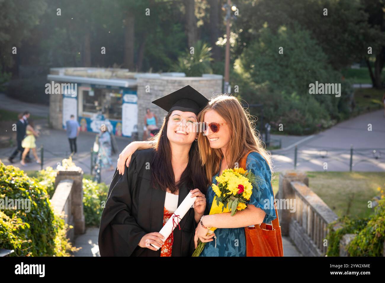 Zwei glückliche Frauen feiern den Abschluss mit einer in Mütze und Kleid mit Diplom und Blumen, und die andere umarmen sie, Bournemouth, Dorset UK Stockfoto