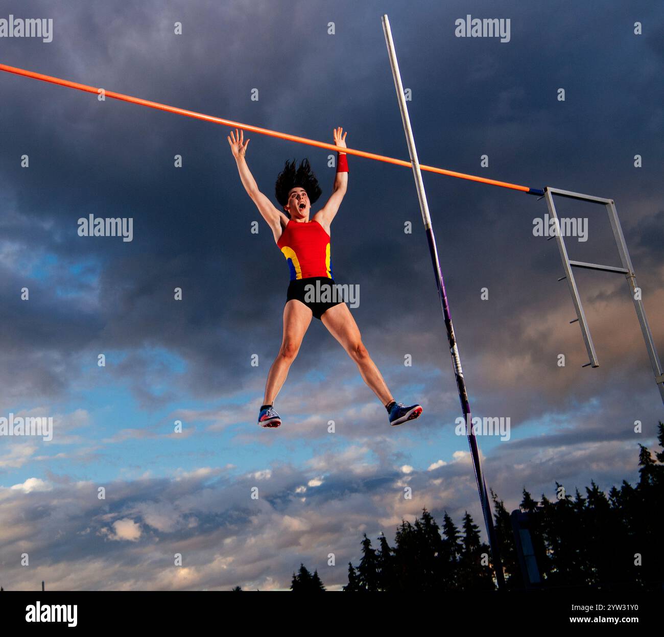 Weiblicher Stabsprung in der Luft bei klarem Sonnenuntergang mit einem hohen Balken gegen teilweise bewölktem Himmel. Stockfoto