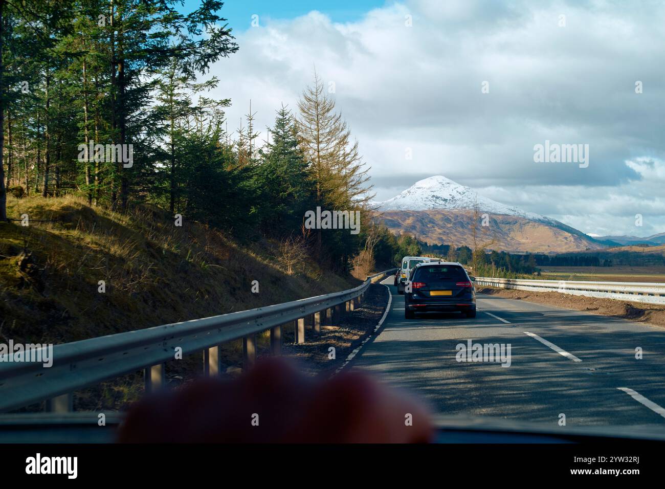 Auto fährt auf einer Straße mit einem schneebedeckten Berg im Hintergrund und Pinien säumen die Route unter einem blauen Himmel mit Wolken, Highlands, Schottland Stockfoto