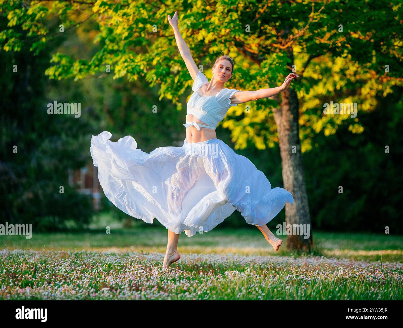 Eine Frau in einem weißen, fließenden Kleid tanzt anmutig auf einem sonnendurchfluteten Feld mit Bäumen im Hintergrund, USA Stockfoto