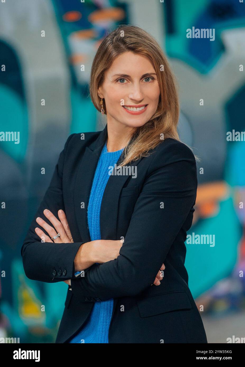 Selbstbewusste Geschäftsfrau in blauem Oberteil und schwarzem Blazer mit überkreuzten Armen vor einer farbenfrohen Graffiti-Wand. Stockfoto