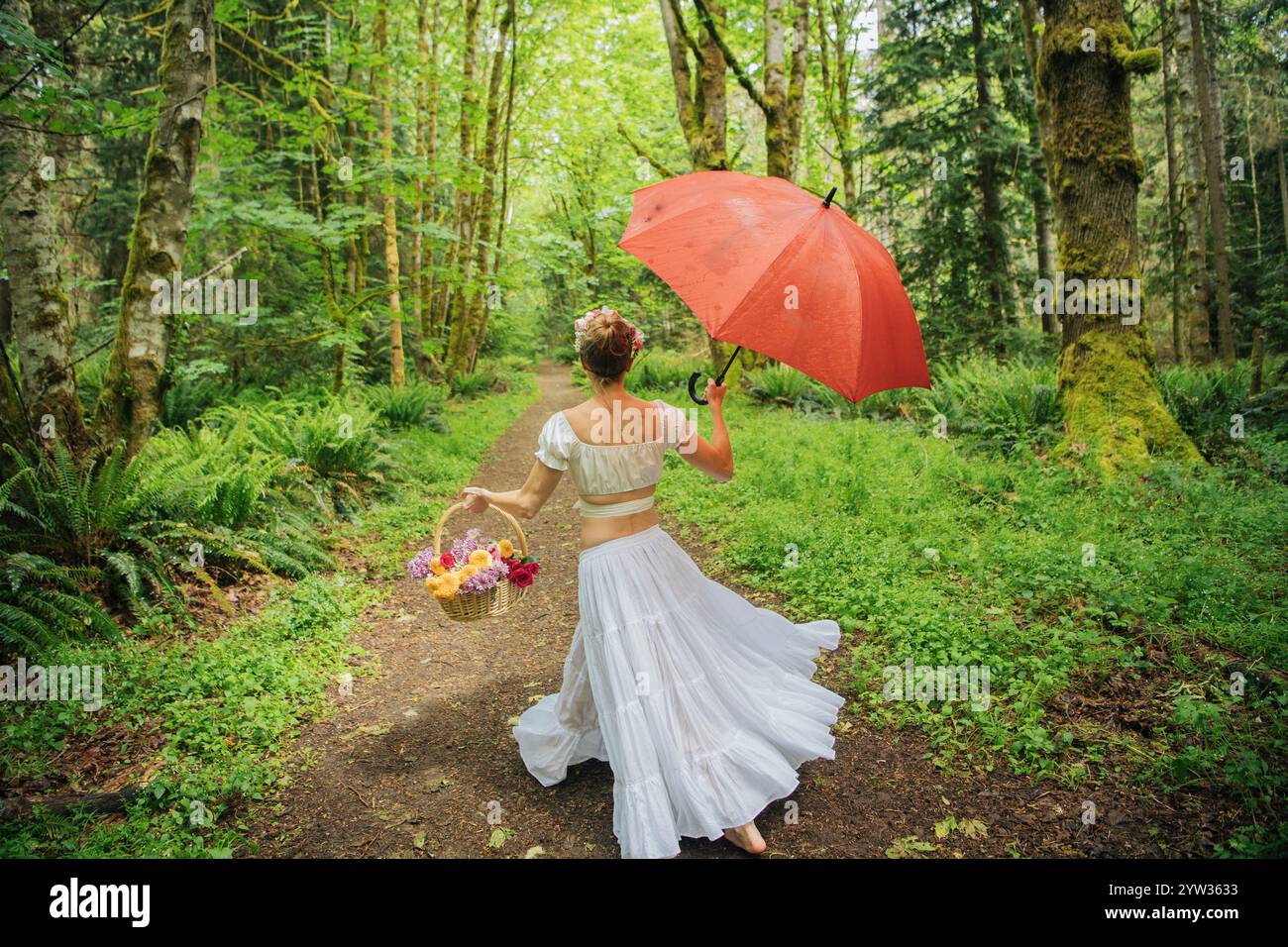 Frau in einem weißen Kleid, die einen roten Regenschirm und einen Korb mit Blumen hält, während sie auf einem Waldweg läuft, USA Stockfoto