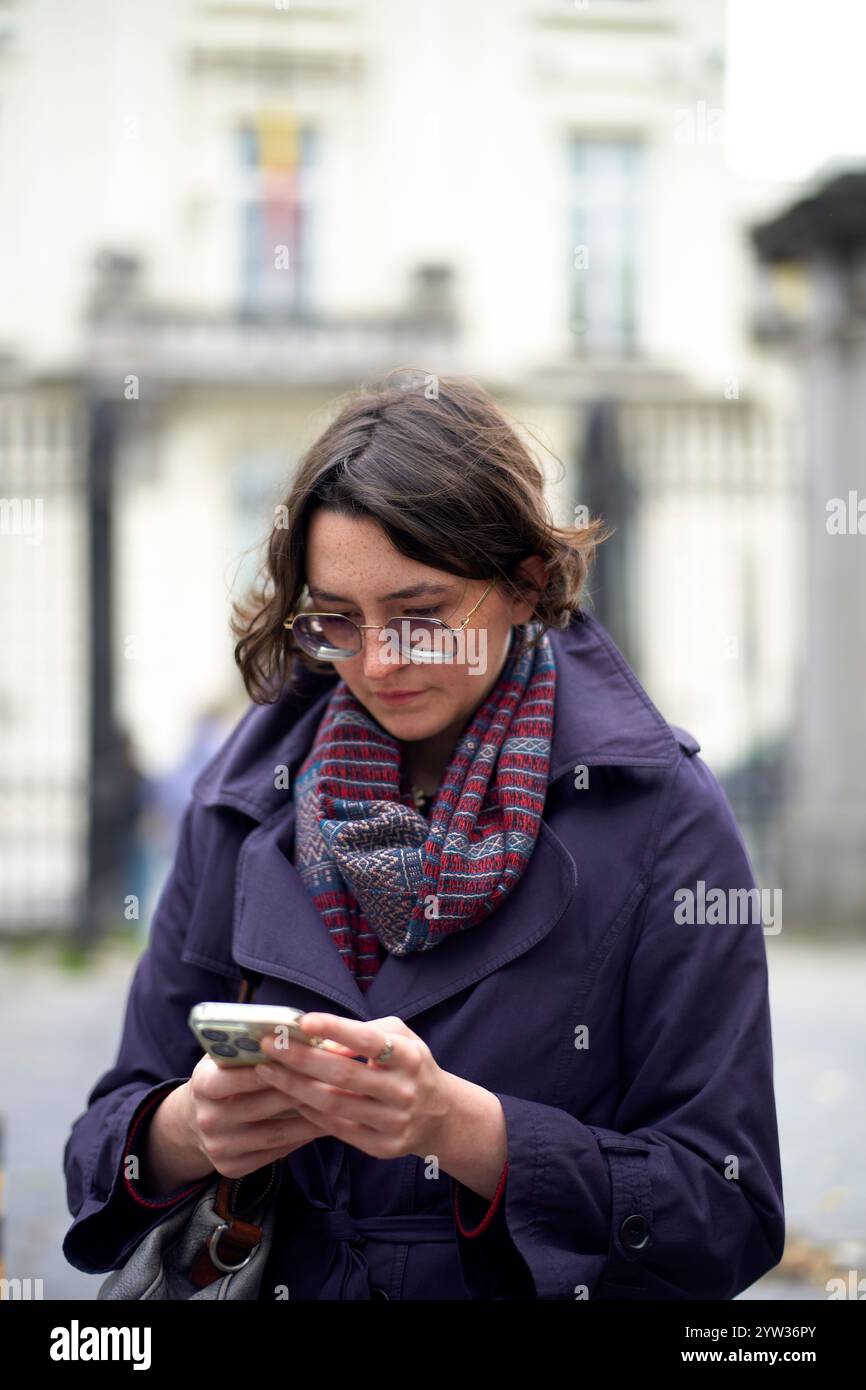 Eine Frau in einem lila Mantel und einem karierten Schal konzentriert sich auf ihr Smartphone mit einem verschwommenen Gebäude im Hintergrund. Stockfoto
