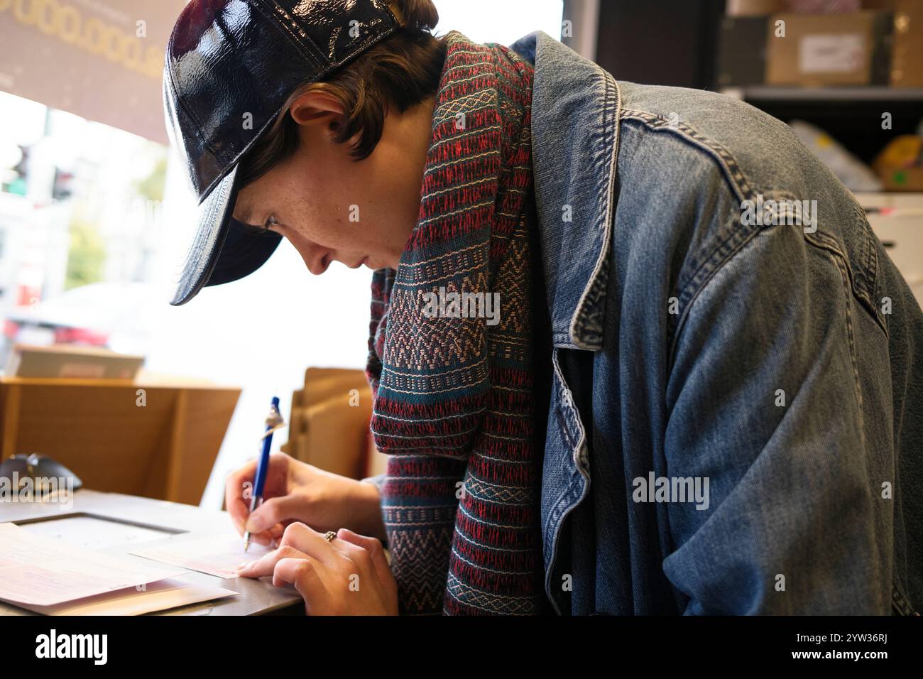 Eine junge Erwachsene Frau in einer Jeansjacke und einer Baseballmütze schreibt an einem Tisch auf ein Blatt Papier und konzentriert sich intensiv. Stockfoto