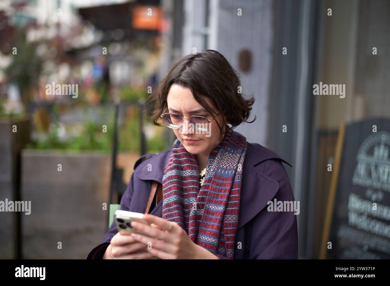 Eine fokussierte Frau in einem lila Schal überprüft ihr Handy in einer Stadtstraße. Stockfoto