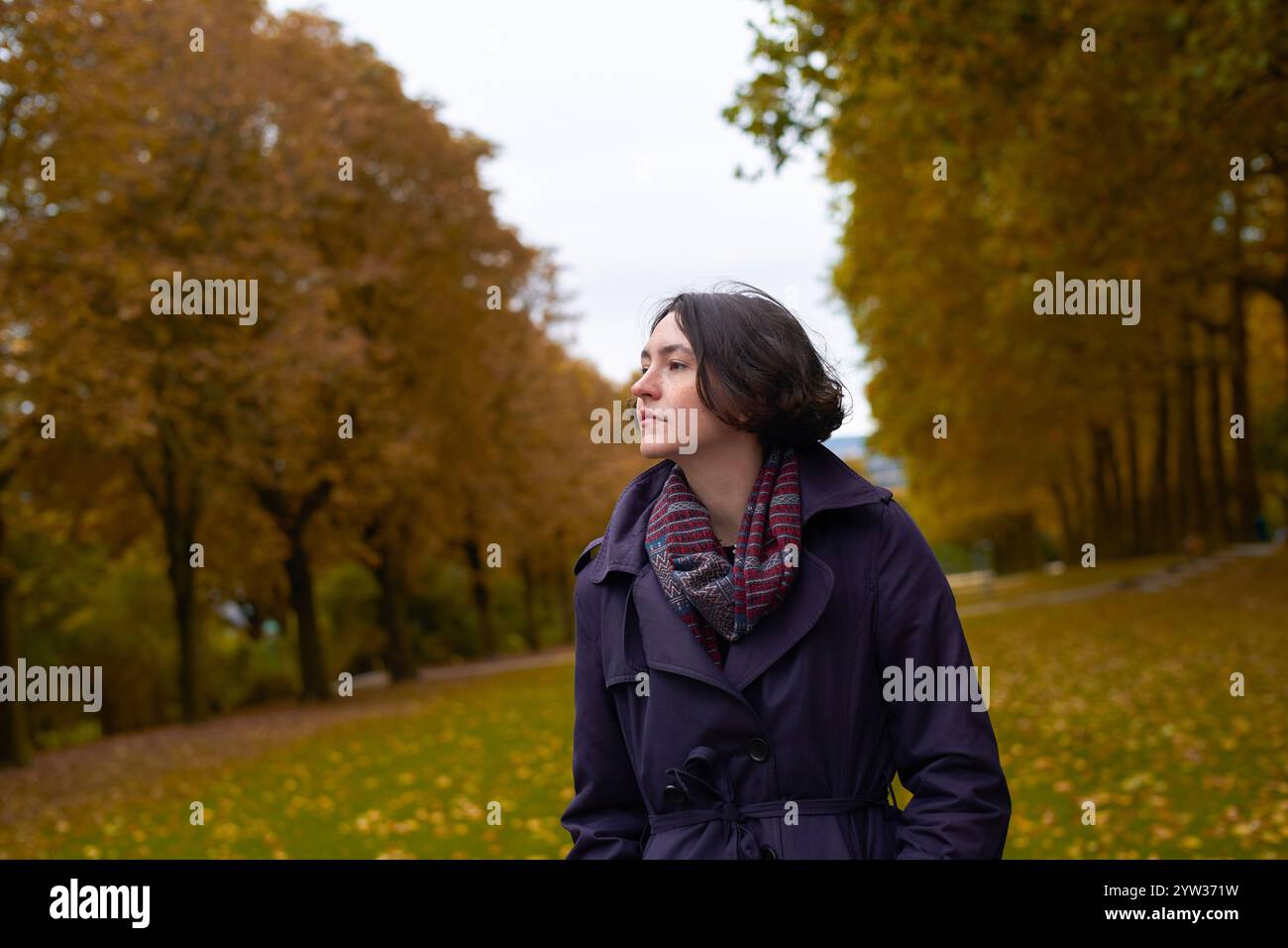 Eine nachdenkliche junge Frau in lila Fell und kariertem Schal schaut zur Seite in einem Park mit goldenen Herbstblättern. Stockfoto