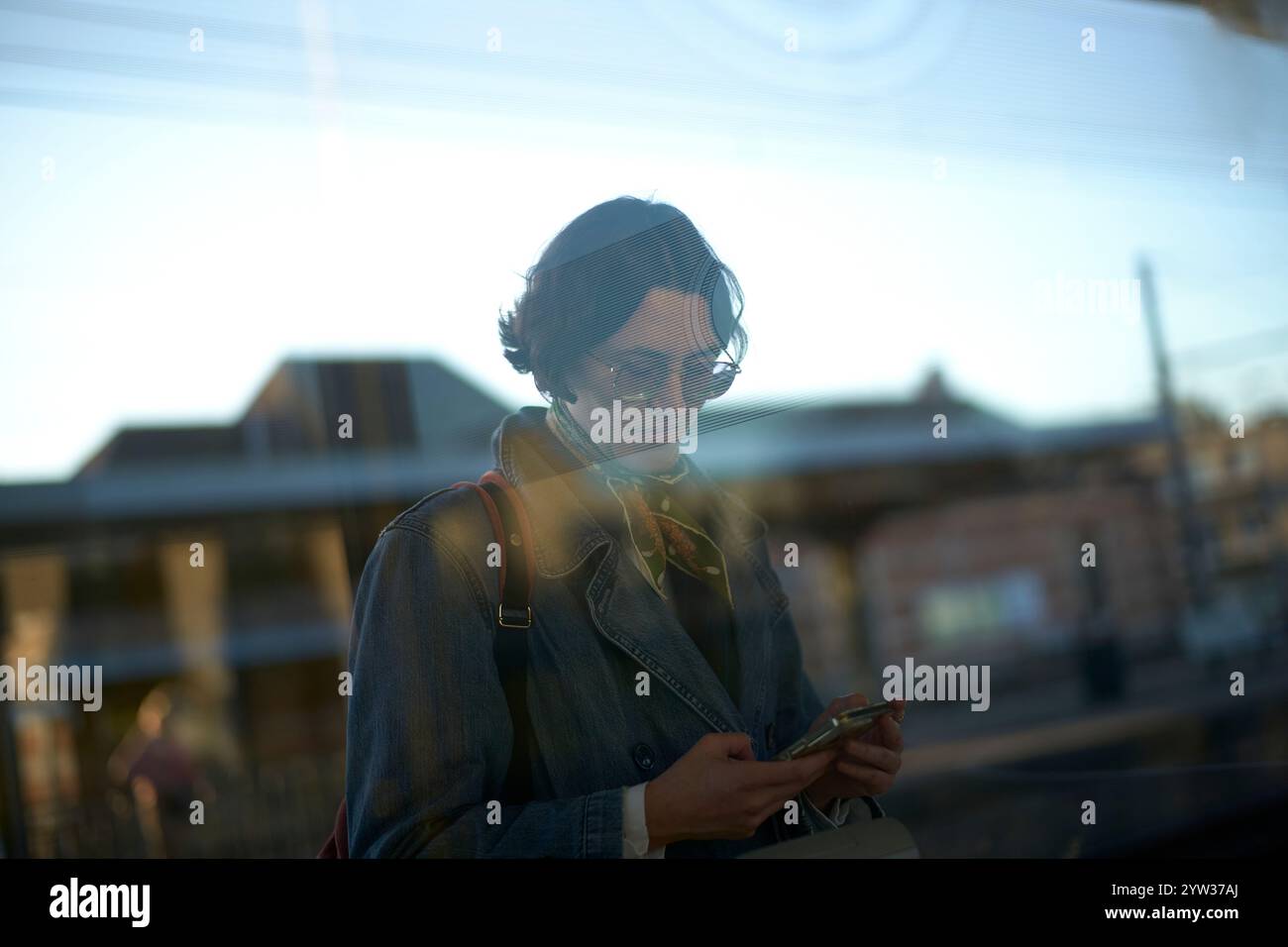 Junge Frau, die ein Smartphone hinter einem Fenster benutzt, mit Reflexionen der städtischen Umgebung, die ihr Bild überlagern. Stockfoto