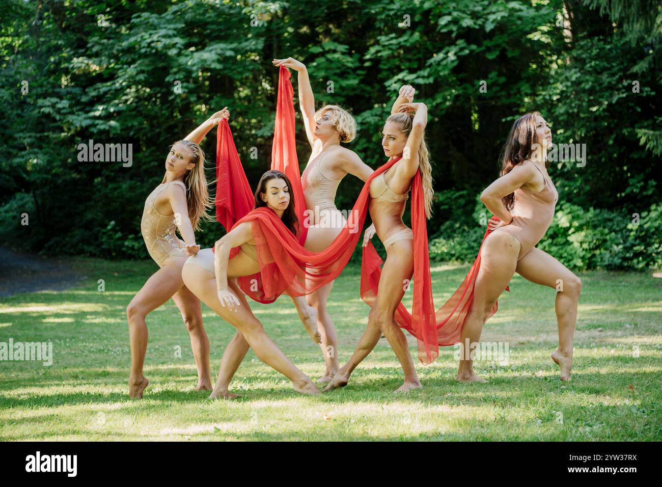 Fünf Tänzer in Bodys spielen einen ausdrucksstarken Tanz mit rotem Stoff in einem sonnendurchfluteten Park, USA Stockfoto