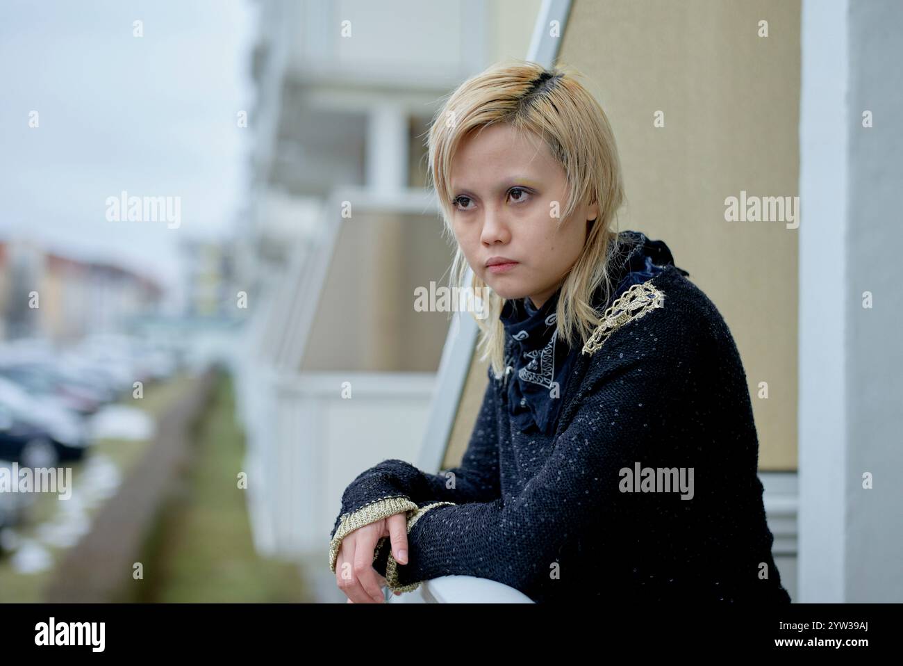 Eine nachdenkliche junge Frau mit blonden Haaren, die draußen auf einem Balkon sitzt und an einem bewölkten Tag nachdenklich aussieht, in Ostdeutschland Stockfoto