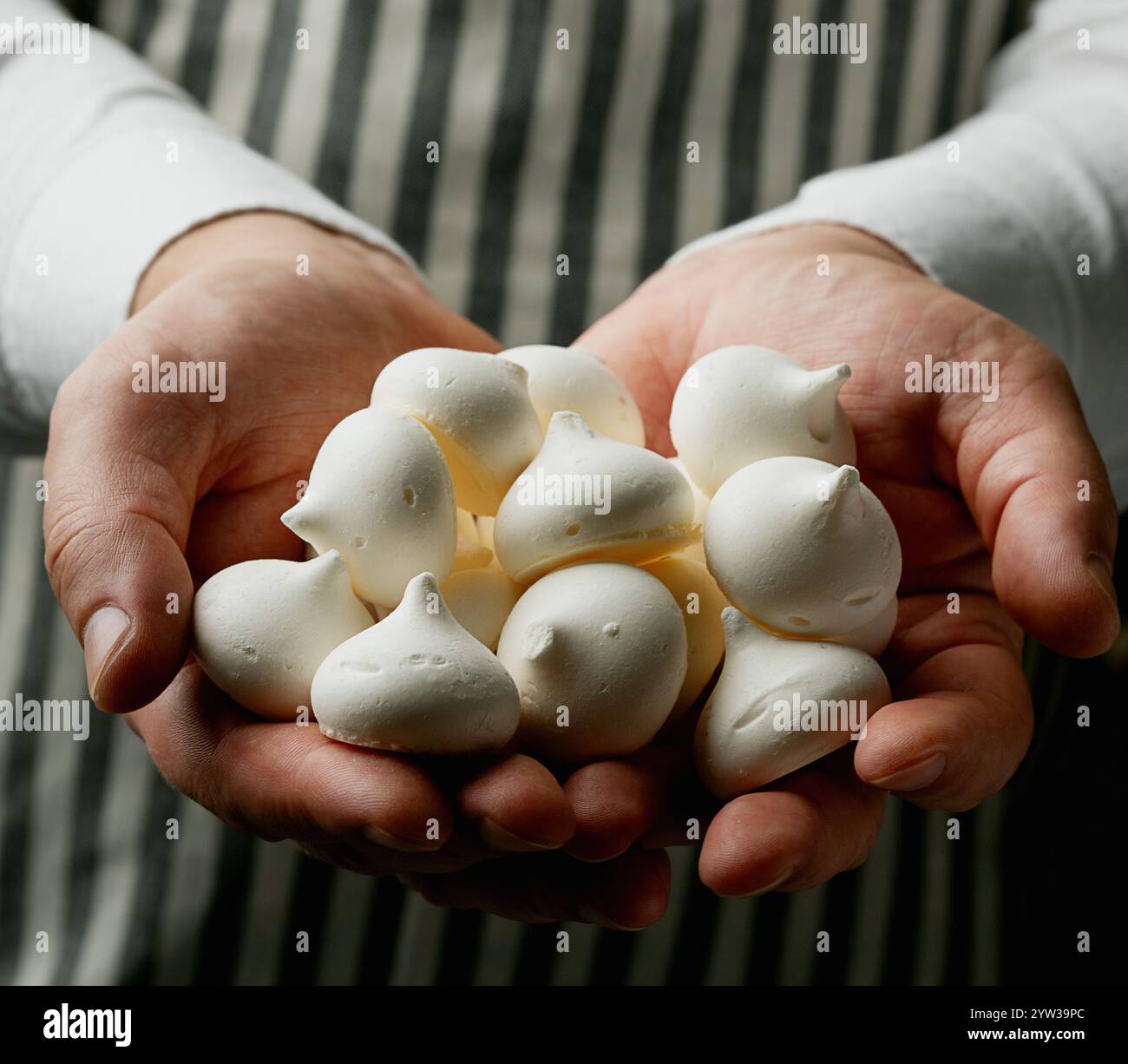 Nahaufnahme von Händen, die kleine weiße Meringue-Kekse halten. Natürliches Licht, gestreifter Schürzenhintergrund für eine minimalistische und elegante kulinarische Szene. Stockfoto