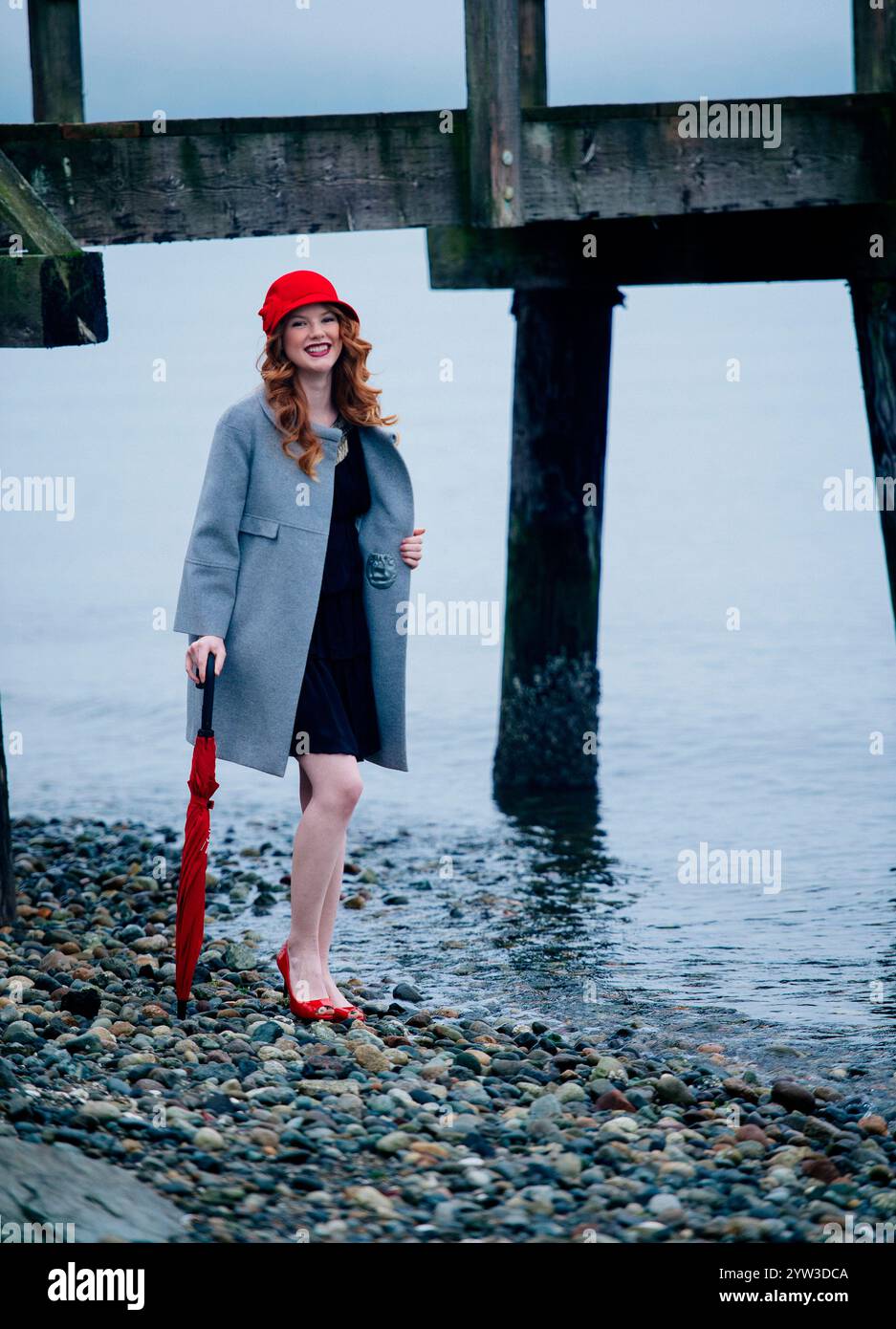 Lächelnde Frau mit rotem Haar, grauem Mantel, roter Baskenmütze und High Heels steht am Meer unter einem hölzernen Pier. Stockfoto