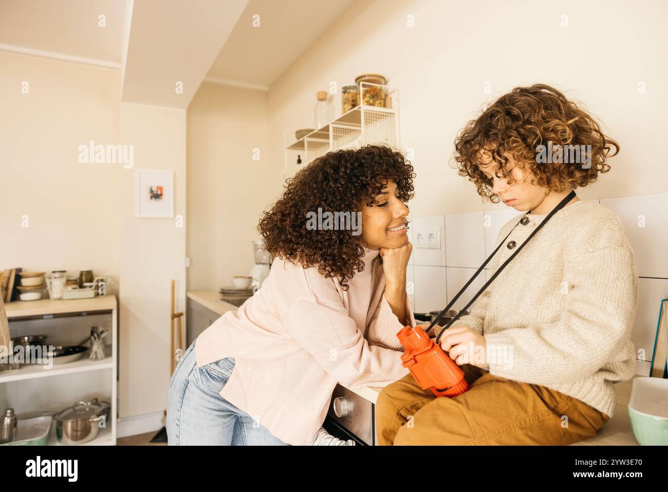 Eine liebevolle Mutter interagiert mit ihrem Kind in der Küche und fördert Wärme und Verbundenheit. Die gemütliche Atmosphäre unterstreicht die starke Familienbindung und das Stockfoto